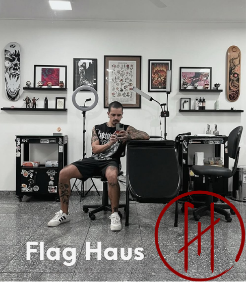 Tatuador sentado em seu espaço de trabalho no estúdio Flag Haus em Vila Mariana, São Paulo, com equipamentos organizados e obras de arte ao fundo