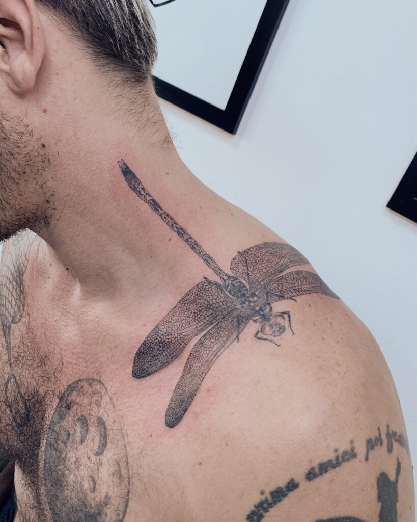 Tatuagem de libélula em fine line e dotwork no ombro, com detalhes de asas realistas, realizada na Flag Haus, Vila Mariana, São Paulo