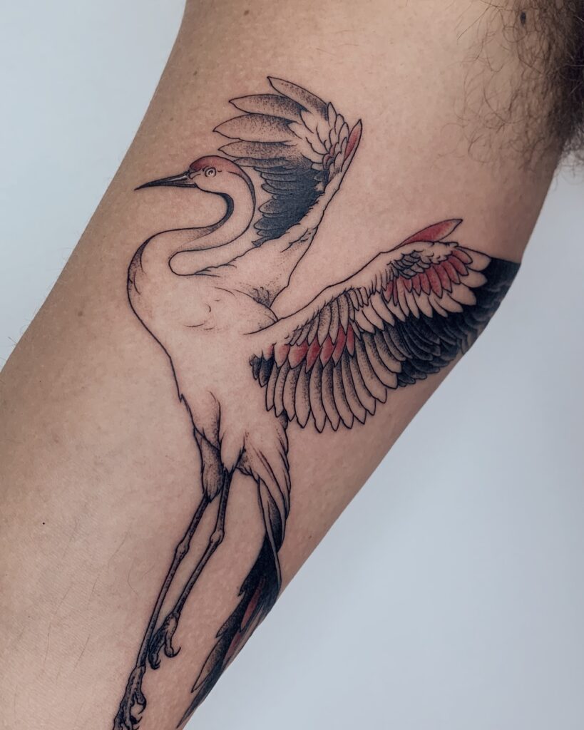 Tatuagem de tsuru japonês em fine line com detalhes em vermelho e preto no braço, realizada na Flag Haus, Vila Mariana, São Paulo