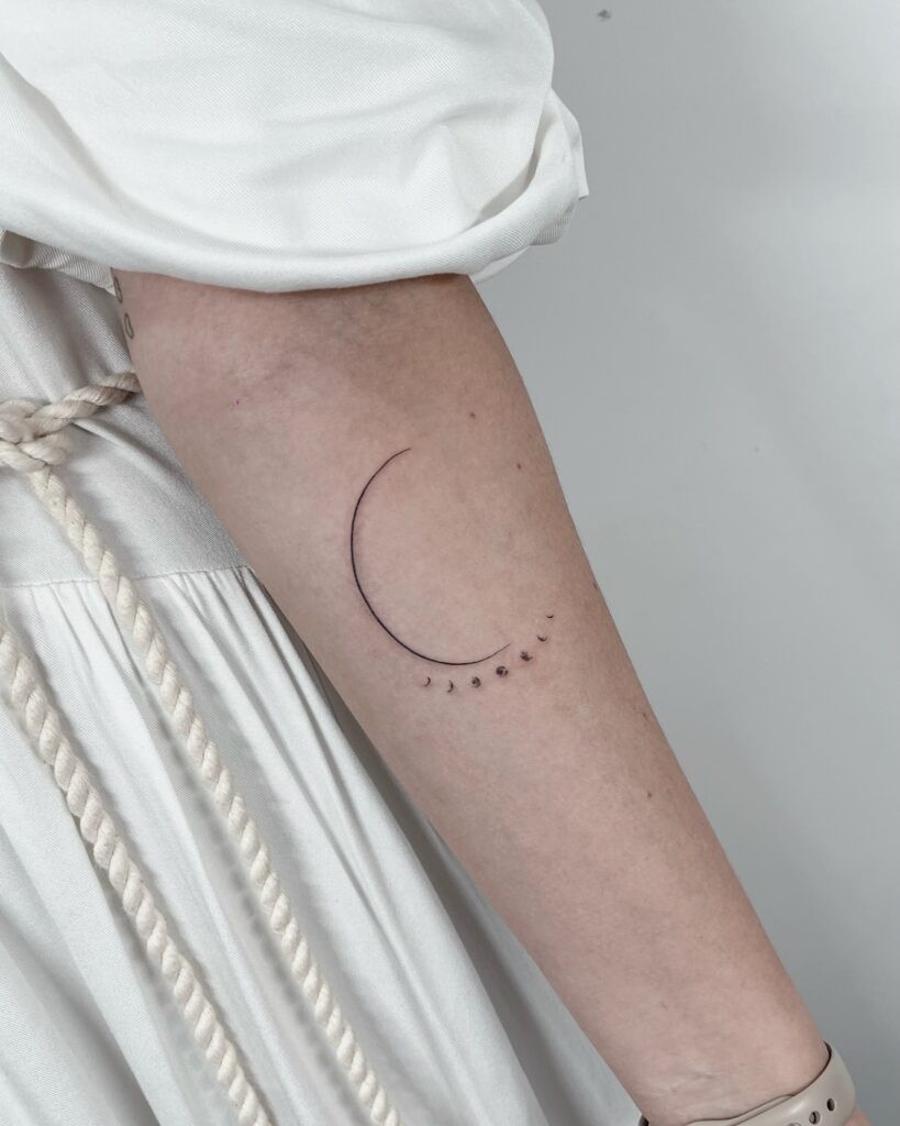 Tatuagem fine line no antebraço: lua crescente em linha única contínua com fases da lua em pontos pequenos, trabalho de traço fino de Julio Bandeiras na Flag Haus