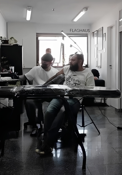 Julio Bandeiras tatuando o braço de um cliente no estúdio Flag Haus, com amigos acompanhando a sessão ao fundo e o letreiro Flag Haus na parede