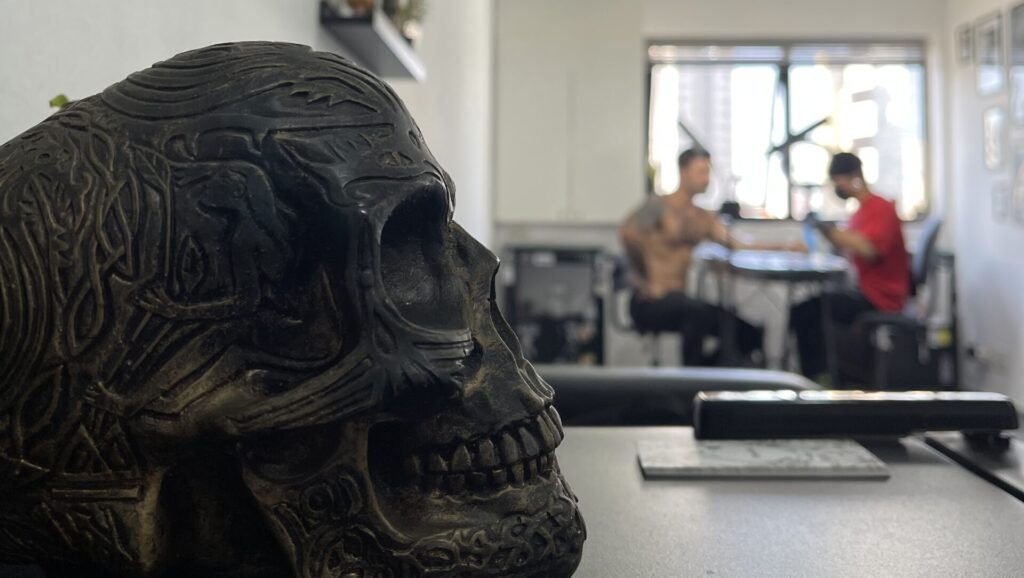 Caveira decorativa entalhada em primeiro plano no estúdio Flag Haus, com Julio Bandeiras tatuando um cliente desfocado ao fundo